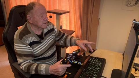 An older man with short brown hair sits at a desk with a monitor. He plays a video game there, controlling a plane with his joysticks just in front of the desk.