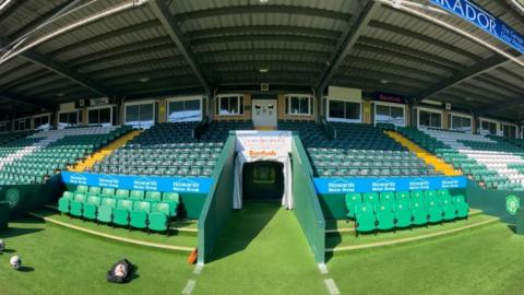 The photo shows the tunnel at Huish Park and the surrounding stand in sunlight. A bag of footballs can be seen on the floor in front of the left hand dugout. 
