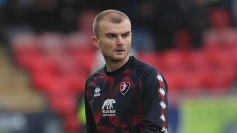 George Miller walking on the pitch in training kit looking over his left shoulder before Cheltenham Town's match against Crewe in January