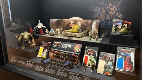 A museum display behind glass of vintage Star Wars toys from 1983's Return of the Jedi film. They include orange, slug-like crime lord Jabba the Hutt who is placed on a toy throne, a brown Rancor monster with claws outstretched, and an alien musical group known as The Max Rebo Band, which features a blue elephant-like creature sitting inside a circular piano-like instrument. There are various other action figures and small vehicles on display, including opened original packaging which once contained such figures.