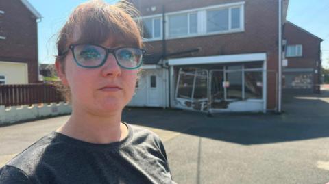 Katy Colquhoun in front of her shop, which has the windows smashed and security fencing around it. She has red hair tied back and a fringe. She wears glasses with a blue frame and sports a grey t-shirt. She is looking at the camera with a serious expression. The grooming parlour appears to occupy the bottom right (as you look at it) section of a huge, relatively modern two-storey detached house, with a white UPVC front door to the left, but it is in fact part of a small row of shops, unseen behind it. It has a large parking area or drive in front.