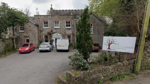 Google Street View image of Ashburton House care home in Ashburton. Three cars and a white van are parked outside the property. It is a cloudy day and there several several trees, shrubs and plants on the outskirts of the site.