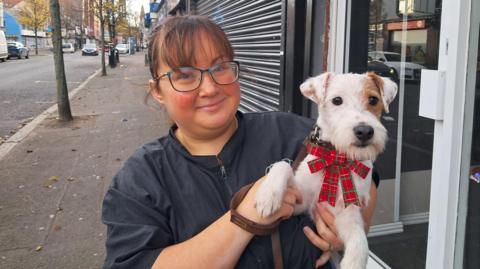 Sarah Teare. She has dark hair tied back with a fringe, wearing glasses and a black zip-up jacket. She is holding a small white dog with brown eye patch and it is wearing a Christmas bow collar. She is standing on a street footpath.