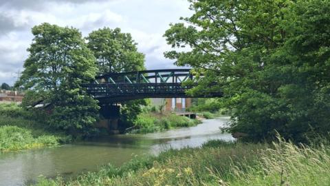 A dark-coloured bridge over a river with trees on both sides and grassy banks. There are brick buildings in the distance and a electricity pylon.