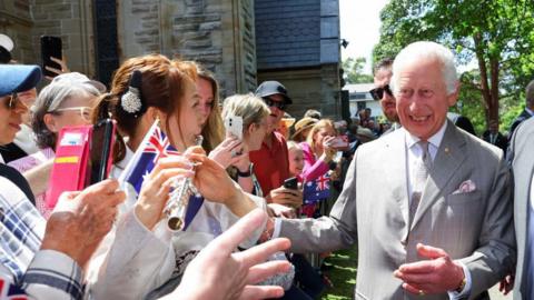 King Charles is on the right side of the image, wearing a silver suit and smiling while looking to a crowd of people directly to his right. He is greeting the crowd who are smiling back at him and taking photos on their phones