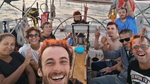 Nine people on a boat sitting together, smiling giving thumbs up and peace signs