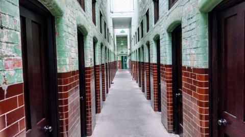 A red and green brick building with a corridor lined by cells.