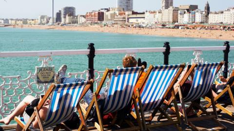 People sit on deck chairs on Brighton Palace Pier overlooking the beach and city of Brighton and Hove.