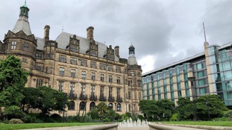 A wide view of the Peace Gardens with a walkway leading toward Sheffield Town Hall. In the foreground, there is a paved path bordered on both sides by low stone walls. At the end of the walkway, a rectangular fountain with multiple small water jets sprays upward in a neat row.
Beyond the fountain, the town hall, a large, historic stone building dominates the scene. The building has an ornate façade with many tall windows, decorative carvings, and multiple chimneys. Its roof features steep slopes and several pointed towers with green-tinted tops, giving it a distinctive, traditional architectural style.