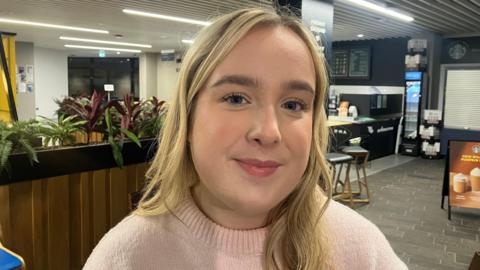 Kiera Pollock is sitting in a cafe. She has wavy blond hair.  She's wearing a light pink jumper. There are some plants behind her and a poster advertising hot chocolate. The cafe is empty.