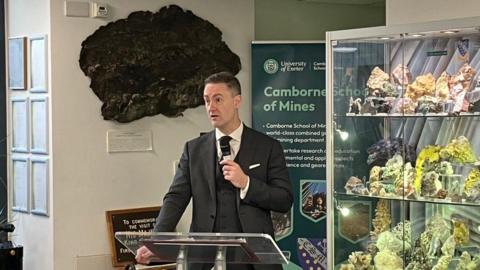 Industry Minister Chris McDonald talks into a microphone while stood at a lectern in front of a cabinet of minerals at the Camborne School of Mines. McDonald is wearing a black suite with a white shirt.