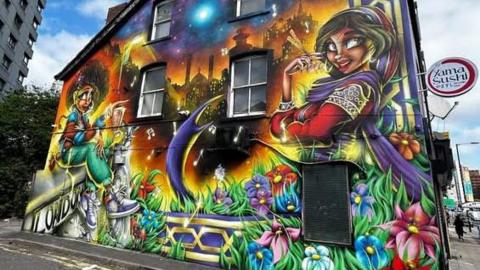 A bold, bright graphic street art wall in a sort of semi-animated style. On the left of the wall, a woman with an afro in green trousers and white trainers sits on a wall. On the right, a woman wearing long sleeved red clothing and a sari, and a purple headscarf, looks at the camera and crosses her arm. Between them is a cityscape at night with music notes floating around, and grass and flowers at the bottom.