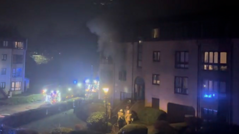 Firefighters respond to a night time building fire, with smoke rising from an upper floor and emergency vehicles illuminating the scene.