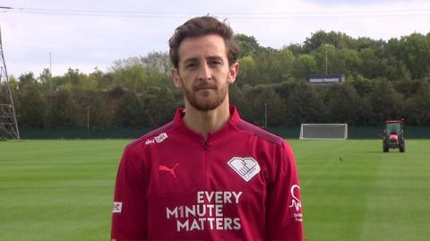 Tom Lockyer, with brown hair and beard, is standing on a football pitch wearing a red football top which says "Every Minute Matters"