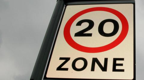 A road sign with the number 20 and the word "zone" in black writing on a white background. There is a red circle around the number, and a cloudy sky in the background.