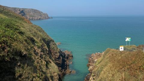 Coastal view of a narrow rocky inlet with clear turquoise water between grassy cliffs, opening out to a calm blue sea beneath a cloudless sky.
