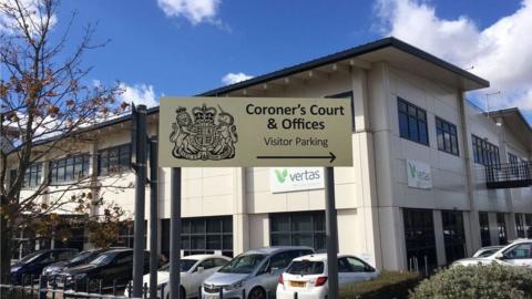 A view of Suffolk Coroner's Court from the outside. It shows the entrance sign with the building sat behind it. Several cars are pictured in the car park directly outside.