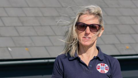 A blonde woman wearing sunglasses is standing in front of a lifeboat rescue station.