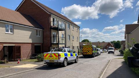 A police car and police forensics van parked outside some houses on Davies Road in Salisbury. Police tape surrounds one of the properties.  