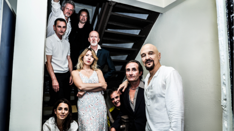 All nine members of the rock band James sit and stand in a stairwell looking into the camera.