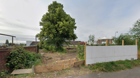 The strip of land has a tree growing out of it and half is fenced off area with the remainder collapsed on the ground. 