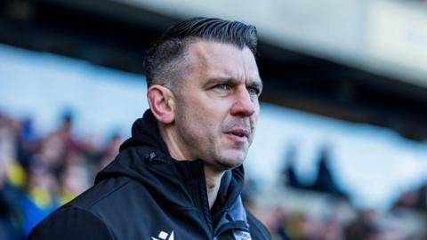 A close up of Matt Bloomfield wearing a black, hooded jacket with a blurred image of Oxford United fans in the background