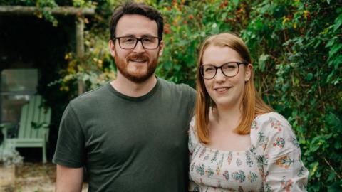 A close up picture of a couple stood outside in a garden. The man on the left has a beard and glasses with a green t-shirt on. The lady is wearing a white dress and also has glasses. There is a green chair in the background.