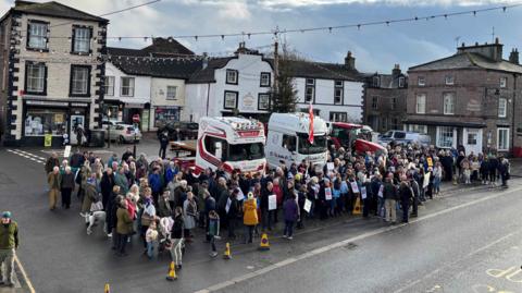 A crowd of more than 200 people with placards. They are standing in a market square in front of two white HGV cabs and a red tractor. There are shops and businesses in the background. 