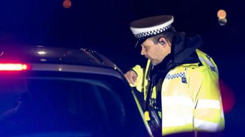A police officers in a high-vis jacket speaking to a driver in a stopped car through the driver's window at night