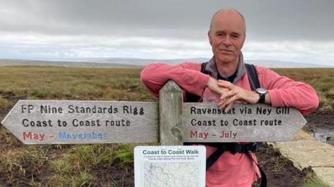 Dr Chris Elliot is wearing a pink jumper and is perched on a wooden sign giving directions in the countryside. 