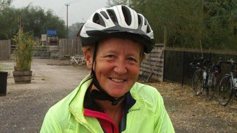 Mary Emerson-Reed smiling into the camera. She is wearing a hi-vis jacket and a cycling. helmet. She is stood in a gravelled area, with trees on the right hand side.