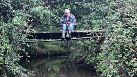 Mike James sitting chained to a small footbridge crossing a stream with foliage on both sides