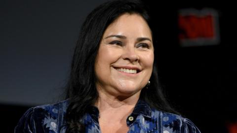 Diana Gabaldon has long black hair and is wearing a blue, patterned shirt. She is smiling as she stands in an auditorium.