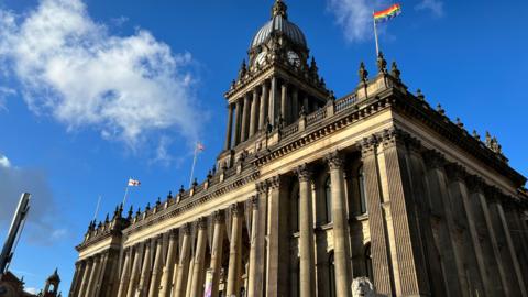 Leeds Town Hall, a grand building with large pillars and a clock tower, with a blue sky and some clouds. 