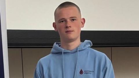 A teenager stands in front  of signage saying Lochlan's Legacy, with further text written underneath. He is wearing a blue top, and has a shaven head.
