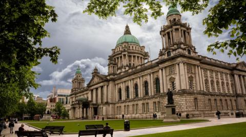 The exterior of Belfast City Hall. It is a large, white Victorian building with blue-domed roofs. There is a large statue of Queen Victoria outside of the main entrance. There is a large statue of an old man to the right of the building, which sits on a neat square of green grass. 