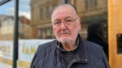 Keith Graham is pictured in a head-and-shoulders shot. He is an older man with receding grey hair and a light grey beard. He is wearing dark-framed glasses and a navy quilted gilet over a dark grey jumper and his expression is serious. He is standing in front of a shop.