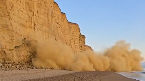 Cliffs to the left of a beach with a large cloud of rubble tumbling onto the beach.