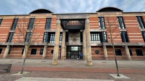 Teesside Crown Court. A large three storey building made from red brick with long narrow black windows. Four large round yellow stone columns support a pyramid shaped glass roof above the front door.