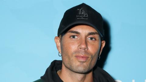 Max George wearing a black cap with the letters L and A intertwined. He is stood in front of a light blue background