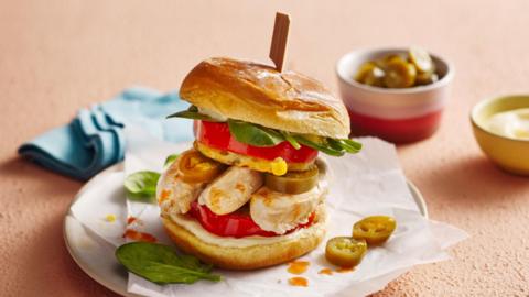 A burger filled with chicken, jalapenos, tomato and leafy greens, sitting on a napkin-laden plate