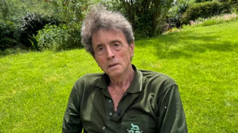 A man with greying wavy hair sits outdoors on a garden chair. He is wearing a dark green polo shirt with a small embroidered crest on the chest. Green lawn, shrubs and mature trees fill the background.