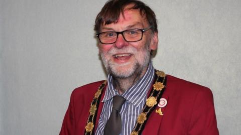 A smiling Stephen Bunney posing for a photograph wearing his gold civic chain. He has a red jacket, a striped shirt and a brown tie. He is wearing glasses and has a grey beard. There are two badges on his lapel.
