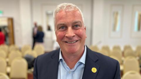 Deputy Andrew Niles - A man with white hair smiling at the camera. He has a blue suit on, with a blue shirt. 