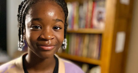 Hephzibah Akinwale who has dark hair in short braids pulled back from her face and is starting to smile. She is wearing a pink and yellow cardigan over a black top. Behind her is a bookshelf full of books.