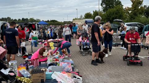 A crowd of people make their way through a car boot sale on a grey day. There is a large amount of toys buyers are stopping to look at as they make their way past.