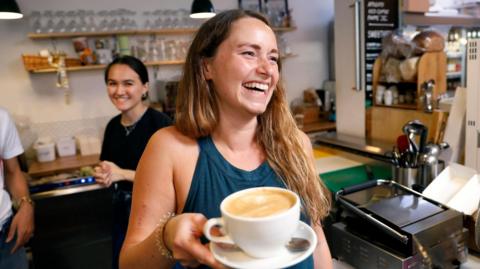 A smiling woman holding a coffee cup in a cafe, with a woman smiling behind her.