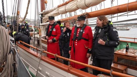 A picture of a number of people stood on a vessel. They are wearing black life vests. Two the people are Chelsea pensioners wearing their red jackets.