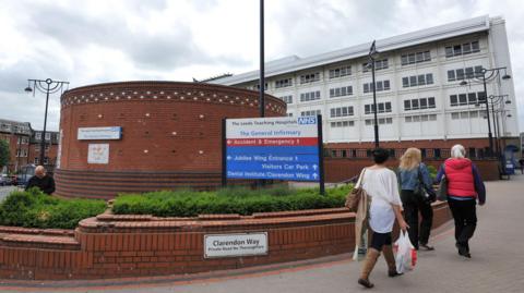 General shot outside Leeds Royal Infirmary. A sign welcoming visitors can be seen whilst three women walk past on the pavement.