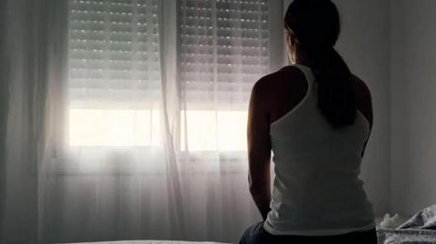 A women sitting on the bed in the dark- bearing a white sleeveless-top with her back towards the camera.